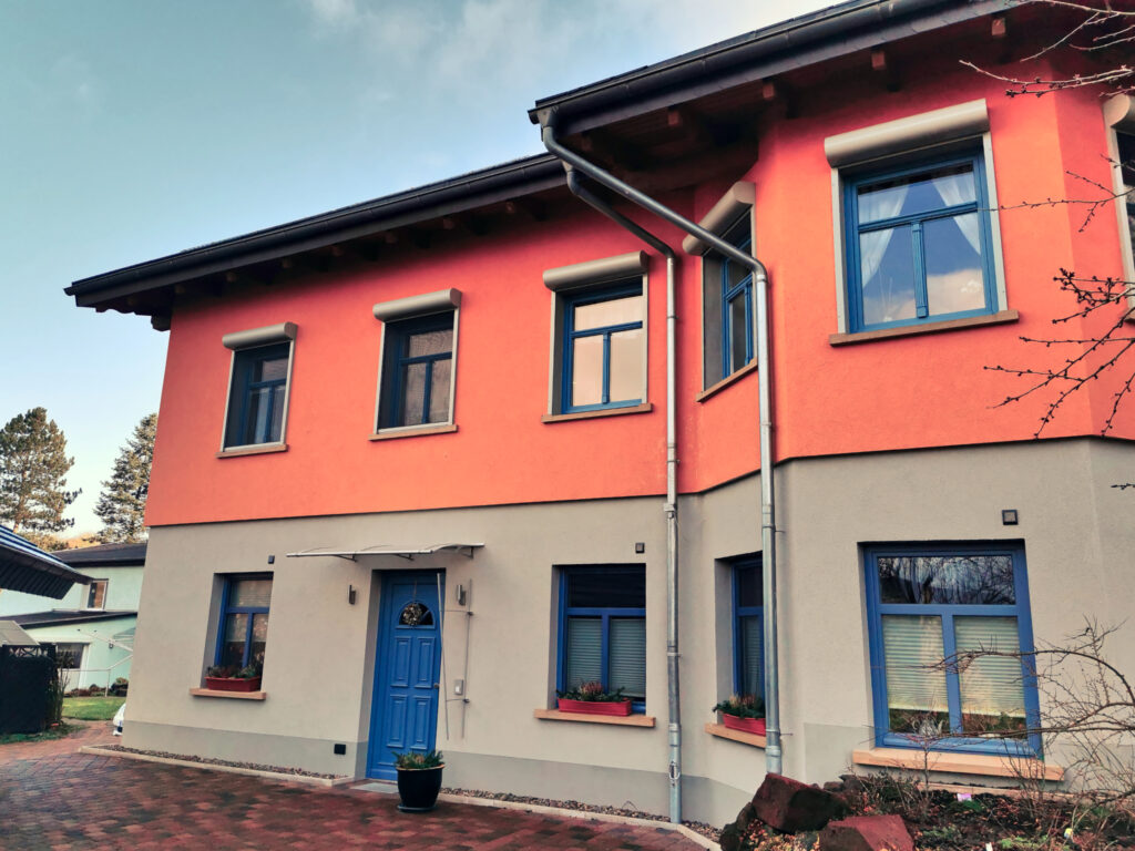 Ein zwei-etagiges Haus, unten grau, oben lachsorange geputzt, mit Fenstern, Giebel und einer blauen Tür.