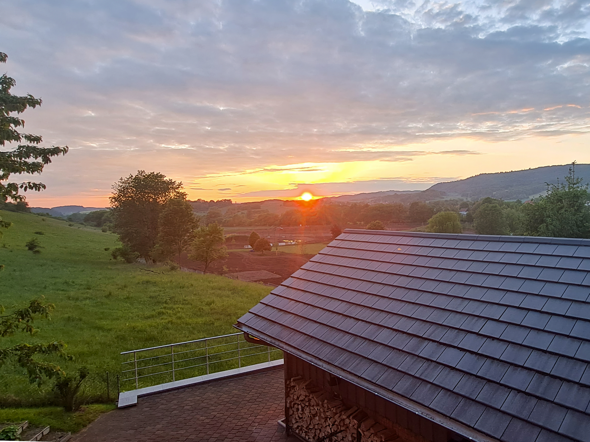 Blick über ein dunkles Ziegeldach zur aufgehenden Sonne hin, hinter dem Haus weite Wiesen und in der Ferne Wald.