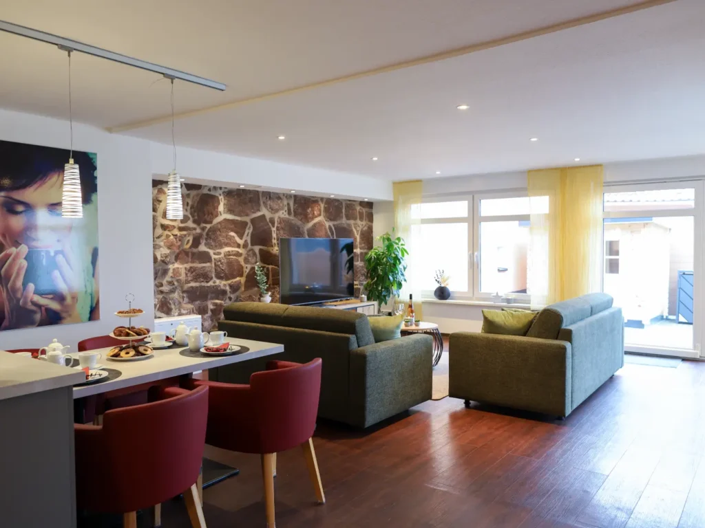 Moderner offener Wohnbereich mit freigelegter Natursteinwand, gemütlicher Sitzecke, Esstisch mit roten Stühlen und Zugang zur Terrasse.