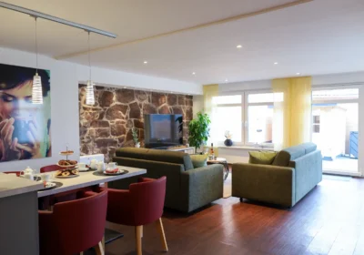 Moderner offener Wohnbereich mit freigelegter Natursteinwand, gemütlicher Sitzecke, Esstisch mit roten Stühlen und Zugang zur Terrasse.