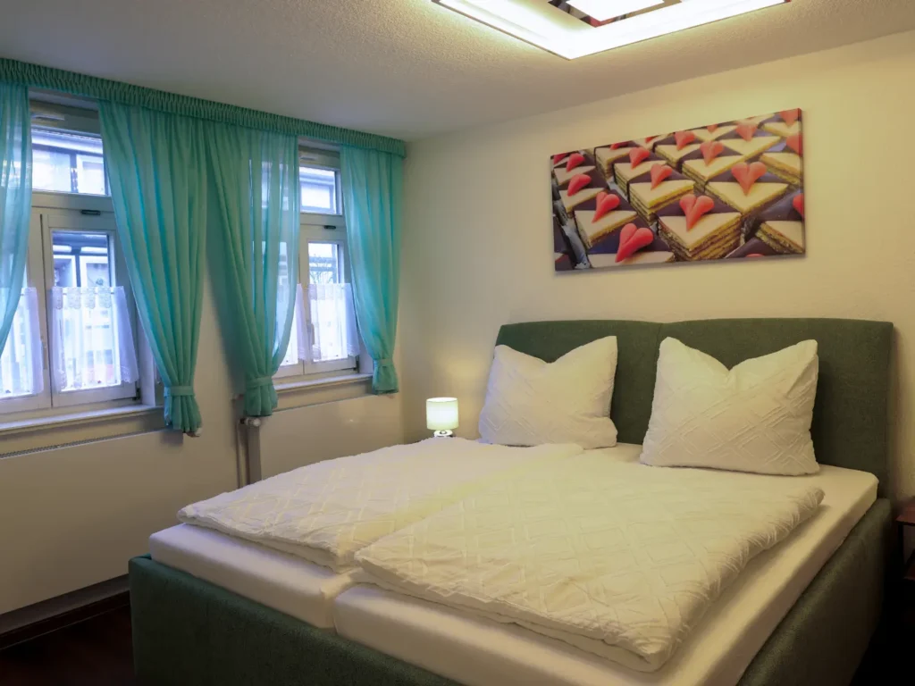 Schlafzimmer mit komfortablem Boxspringbett, mintgrünen Vorhängen und Wandbild mit Törtchen-Motiv passend zur Historie.