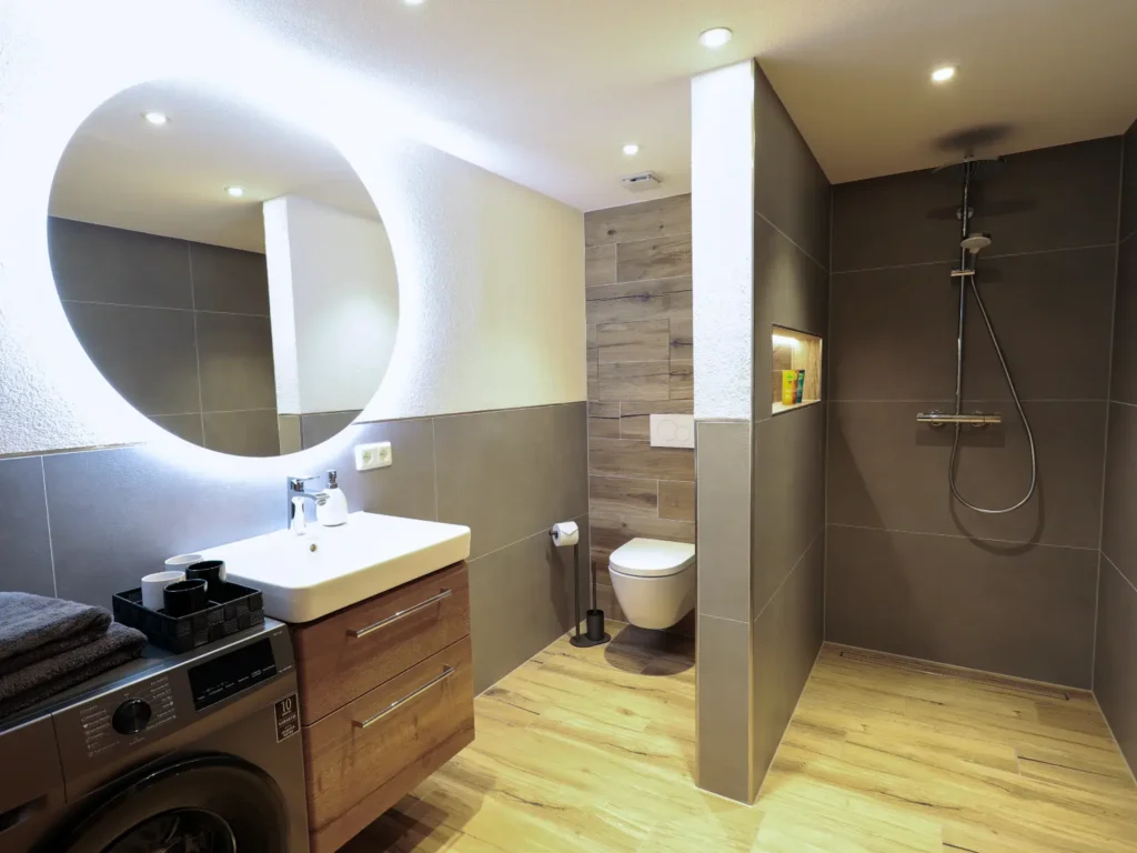 Hochwertiges Badezimmer mit bodengleicher Regendusche, großem runden LED-Spiegel, Waschtisch in Holzoptik und Waschmaschine.