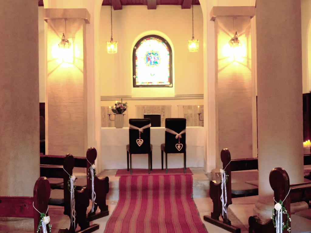 Innenraum der Krypta, hergerichtet zur Trauung: Mittelgang mit rot-rosa Läufer, dunkle Holzbänke mit weißen Schleifen, zwei Stühle mit Holzherzen vor dem Altar, dahinter ein buntes Glasfenster.