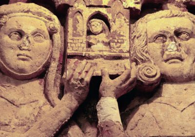 Steinernes Stifterrelief in der Krypta der Neanderklinik in Ilfeld: zwei Köpfe halten ein kleines Kirchenmodell mit Nische und winziger Figur. Der Stein ist hellbeige, in den Vertiefungen Reste rötlicher Fassung.