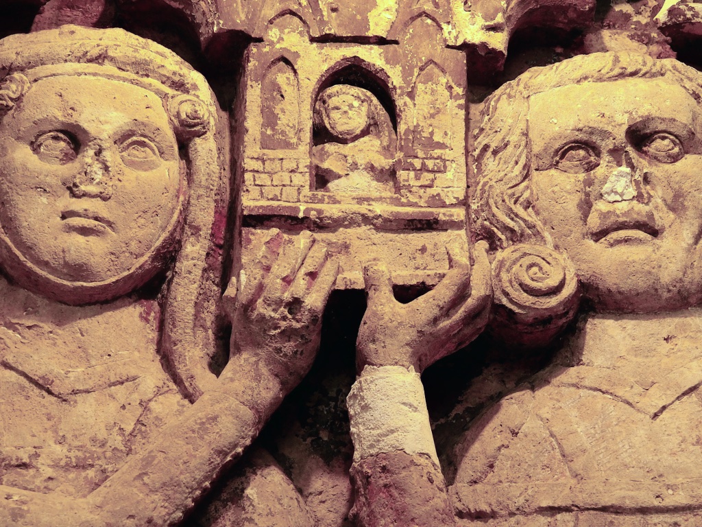 Steinernes Stifterrelief in der Krypta der Neanderklinik in Ilfeld: zwei Köpfe halten ein kleines Kirchenmodell mit Nische und winziger Figur. Der Stein ist hellbeige, in den Vertiefungen Reste rötlicher Fassung.