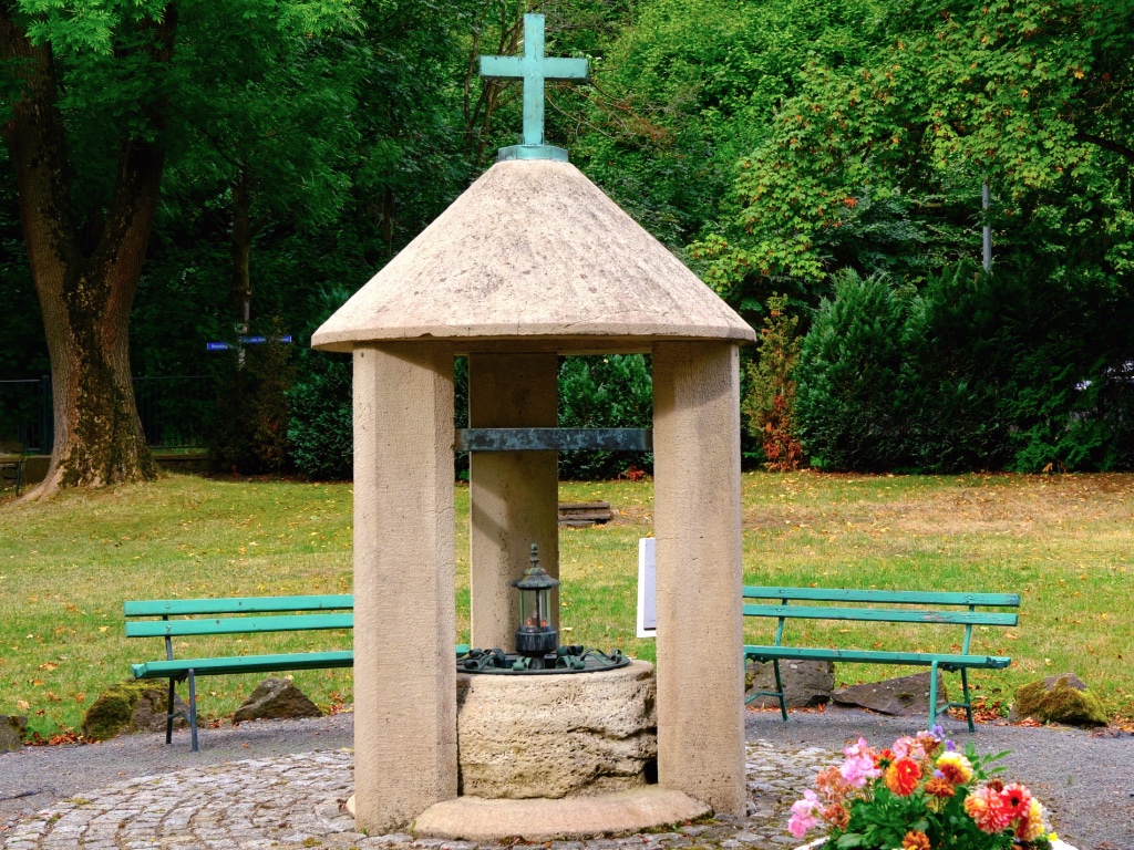 Steinerner Pavillon mit Kreuz auf dem Dach im Park der Neanderklinik in Ilfeld. Unter dem Dach steht eine Laterne auf einem runden Sockel, links und rechts grüne Bänke, vorn Dahlien.