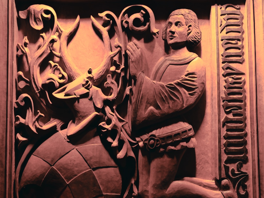 Holzrelief in der Krypta der Neanderklinik: Ein Mann im mittelalterlichen Gewand kniet im Gebet vor einem Wappen mit rautenförmigem Schild, Helm und Geweih. Rechts eine senkrechte Inschrift in gotischer Schrift.