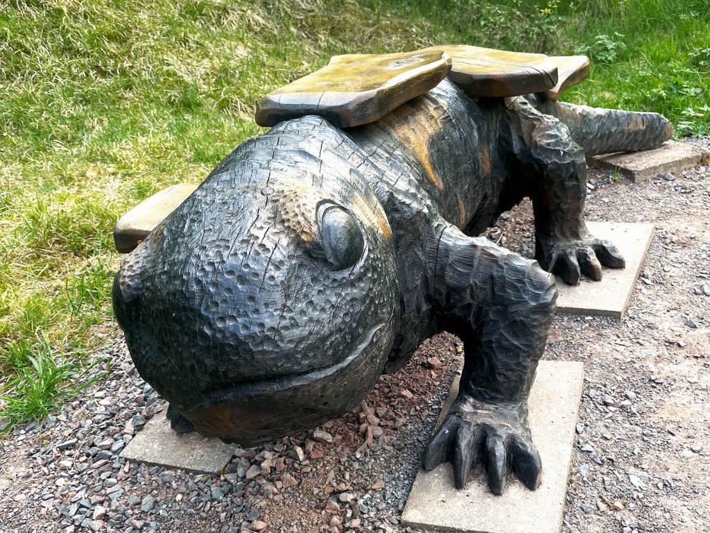 Große Holzskulptur eines Feuersalamanders als Sitzbank am Wanderweg. Der Körper ist dunkelbraun mit hellen Flecken an Kopf und Rücken, auf dem Rücken liegen glatte Holzbretter zum Sitzen. Die Bank steht auf Steinplatten vor einem begrünten Hang.