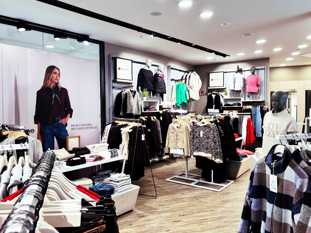 Innenraum Mode Koch Nordhausen mit Street-One-Wandmotiv „Rich Colours. Relaxed Attitude.“, Kleiderstangen, Schaufensterpuppen und Sweatshirt mit Aufschrift „COZY“.